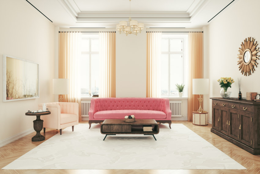 Amplia sala de estar con sofá rosa de un cojín, ventanas con moqueta, lámparas de pie y lámpara de araña